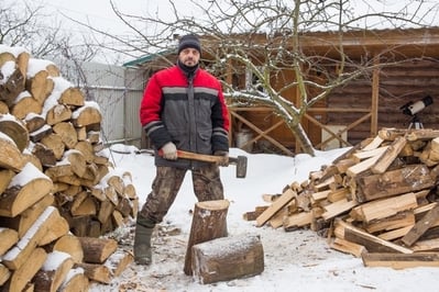 A man stands next to a pile of chopped wood in the winter while holding an axe.